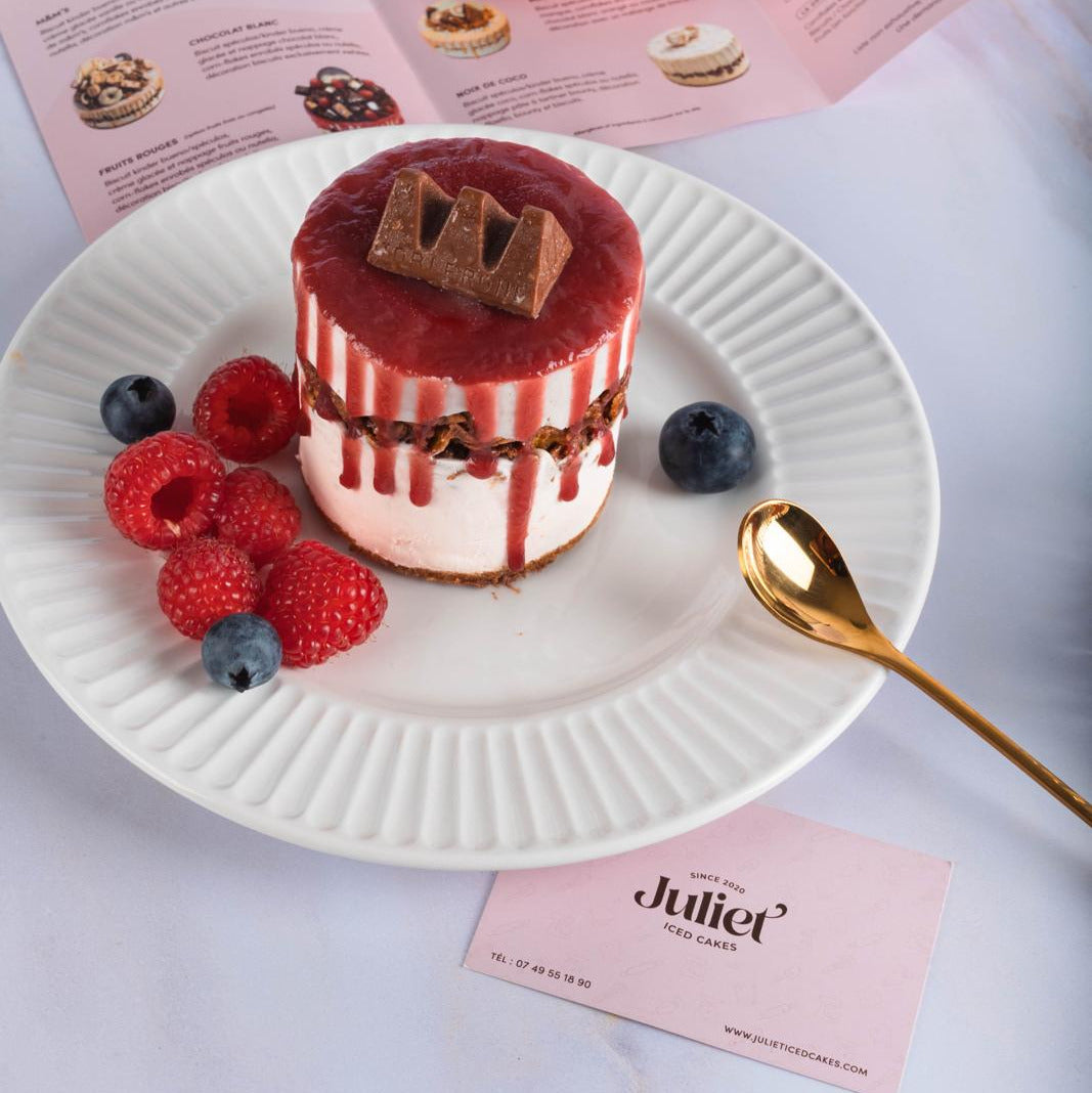 Photo d'un Individuel aux fruits rouges dans une assiette blanche, une cuillère dorée dessus et des flyers et une carte de visite de Juliet Iced Cakes autour.